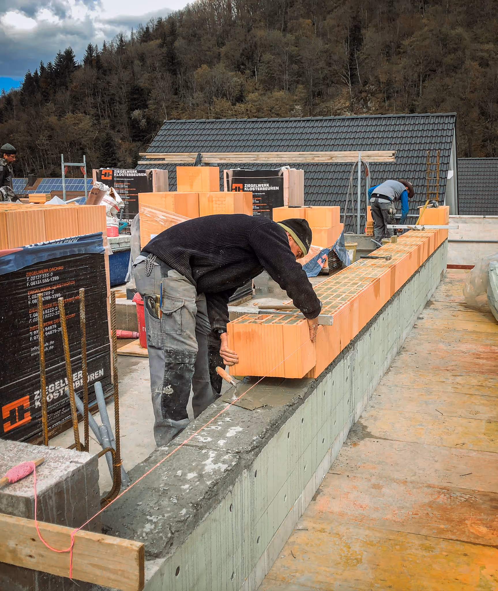 Ein Maurer setzt einen Ziegelstein auf eine frisch betonierte Mauer, im Hintergrund weitere Arbeiter bei der Arbeit.