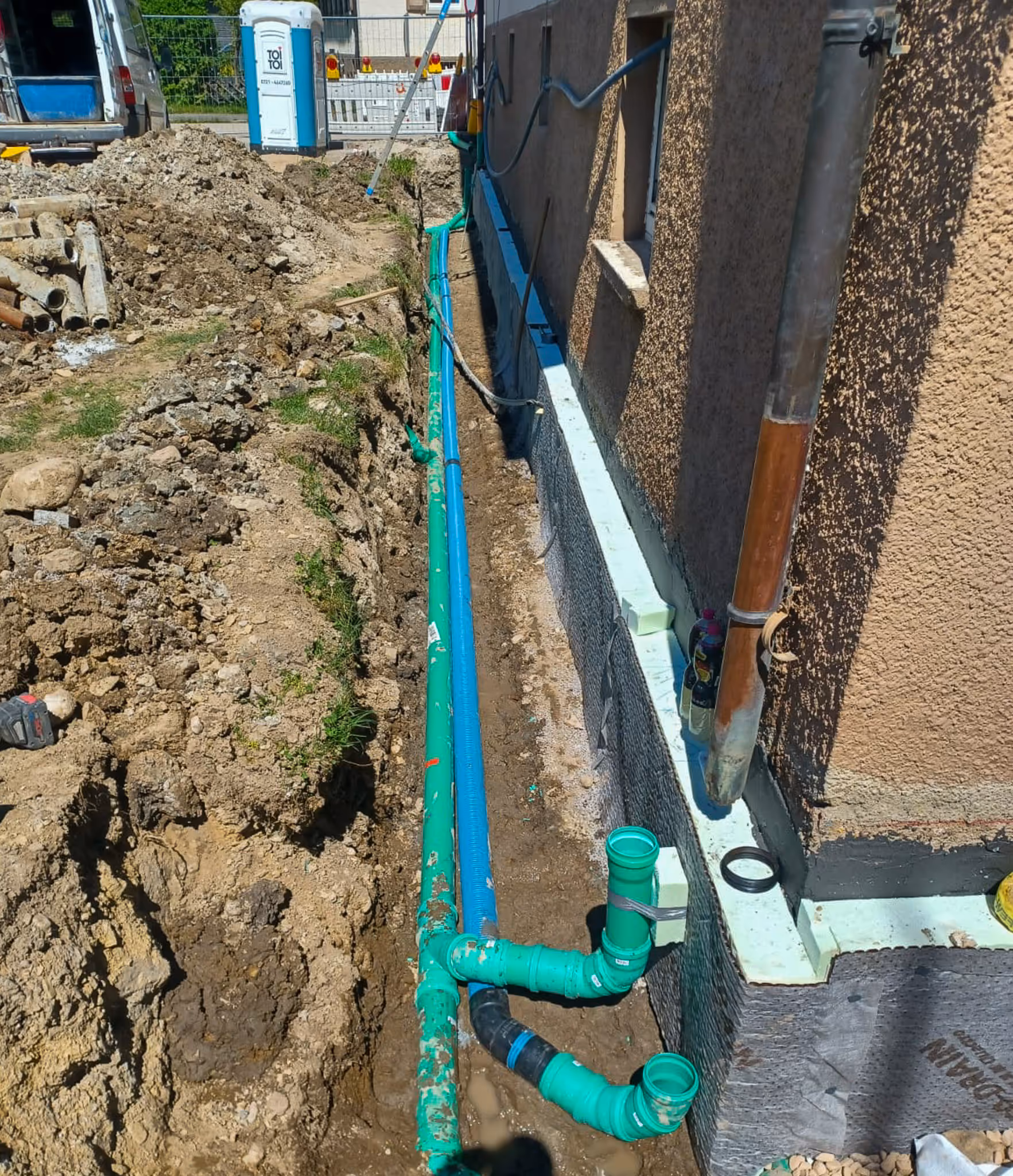 Graben entlang eines Hauses mit neu verlegten grünen Abwasserrohren und Baustellenzubehör.