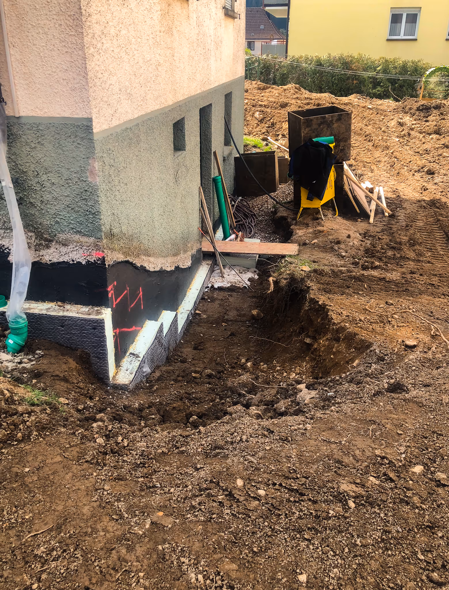 Fundament eines Hauses mit schwarzer Abdichtung, umgeben von Erdreich und Baustellenmaterial.