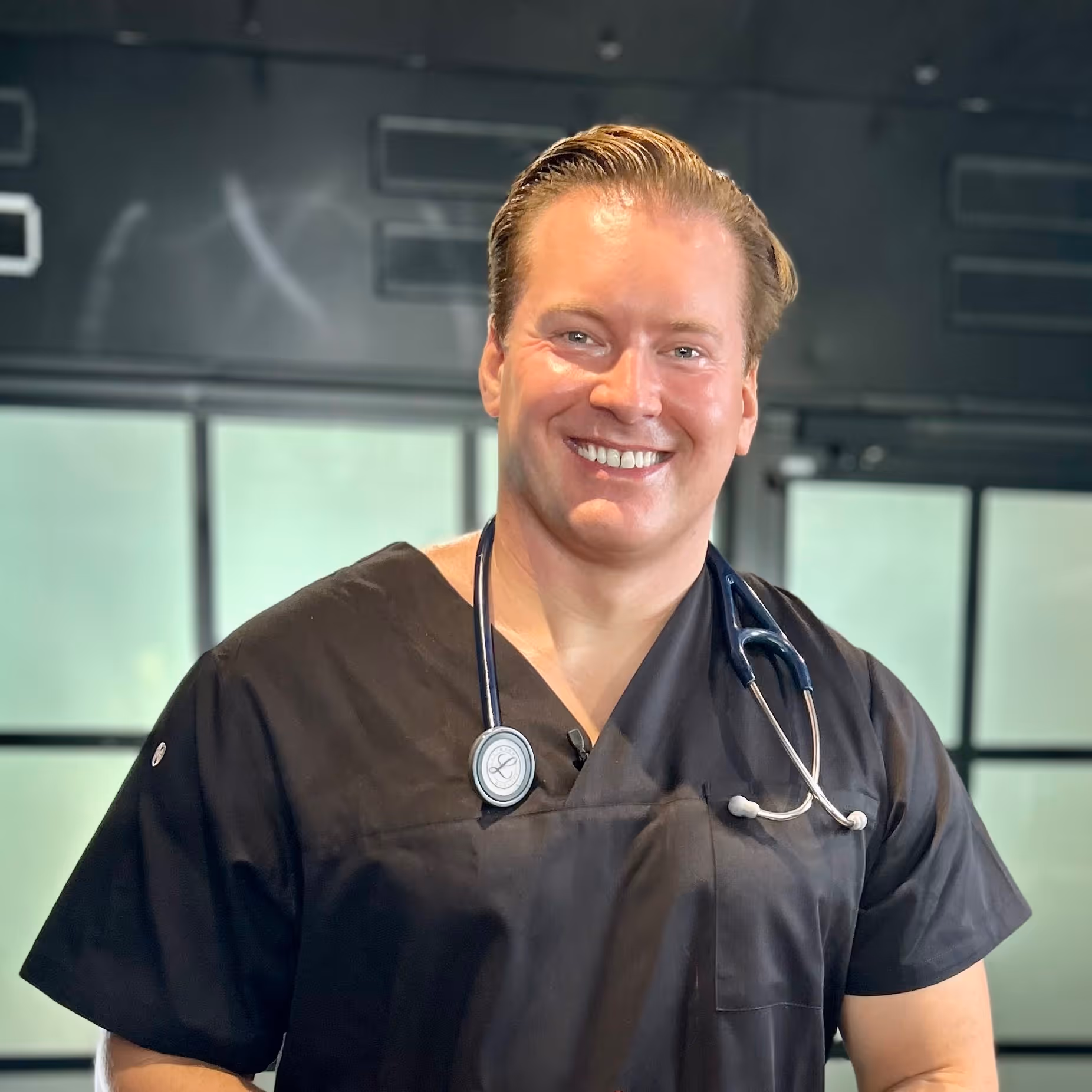 Leende man iklädd svart medicinsk uniform med stetoskop runt halsen i ett modernt rum.