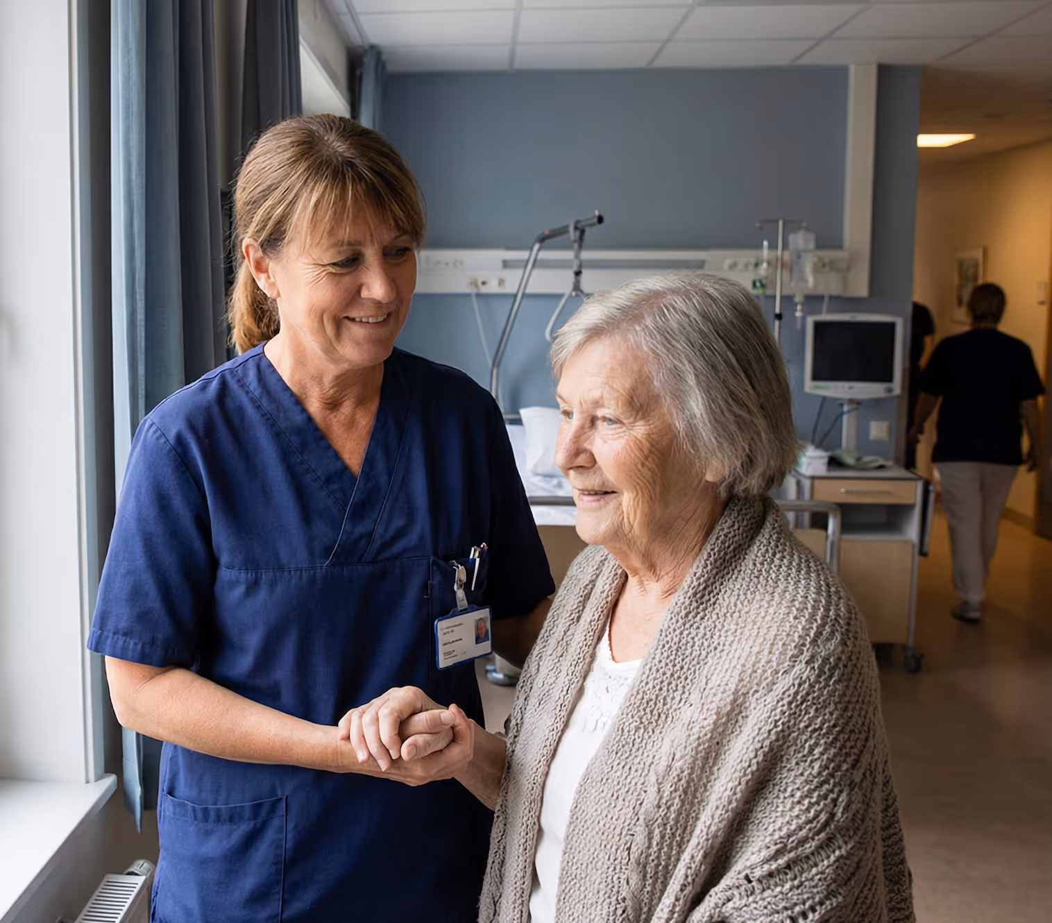 Sjuksköterska som håller äldres hand och ler i ett sjukhusrum.