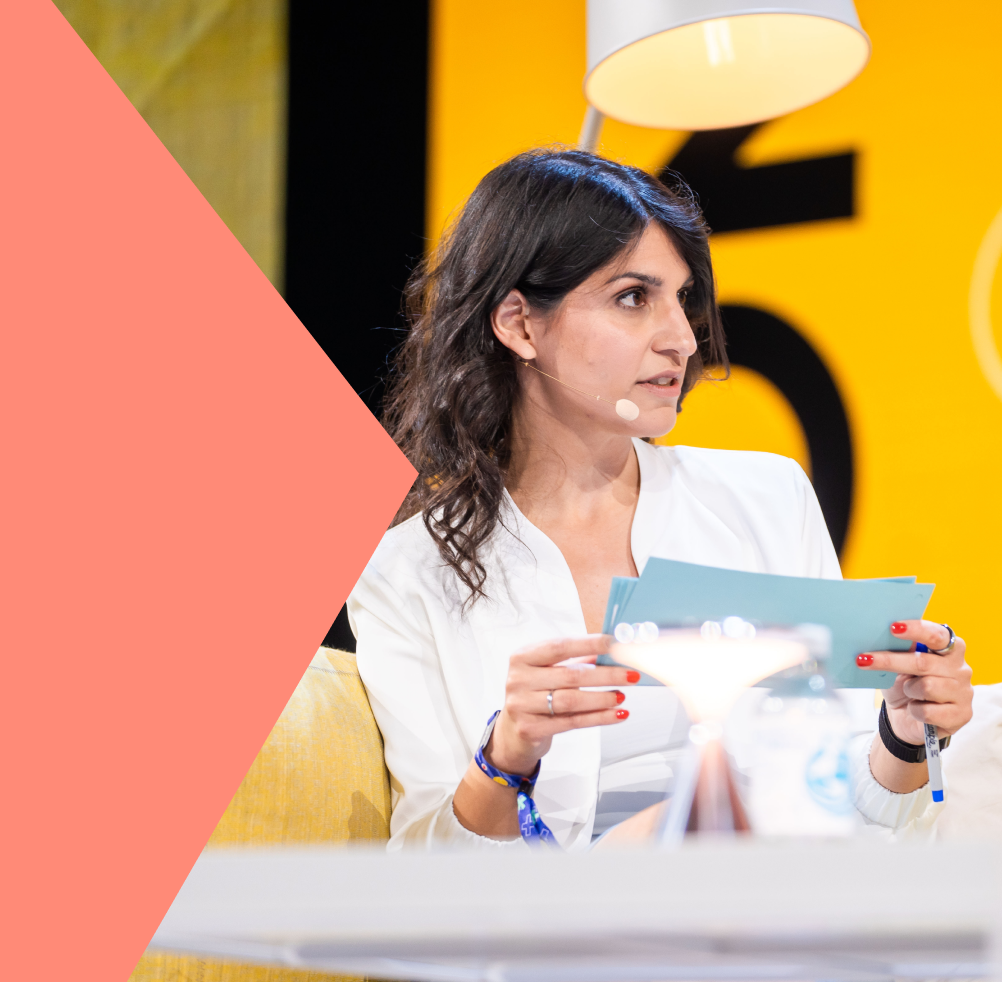 Zamina Ahmad spricht bei einer Veranstaltung mit Mikrofon am Kopf, hält blaue Karten in der Hand vor gelbem Hintergrund.