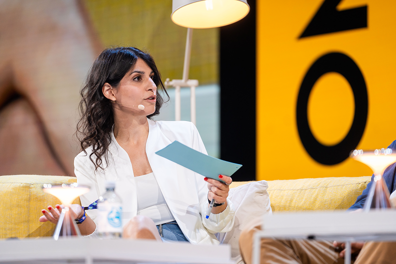 Zamina Ahmad mit weißer Jacke spricht und hält ein hellblaues Blatt Papier während einer Podiumsdiskussion.
