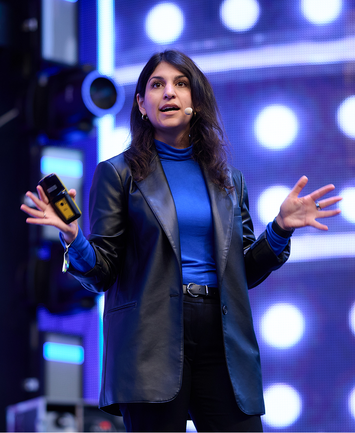 Zamina Ahmad mit dunkelbraunen langen Haaren und Lederjacke spricht auf einer Bühne mit Mikrofon am Ohr, im Hintergrund leuchtende bunte Lichter.