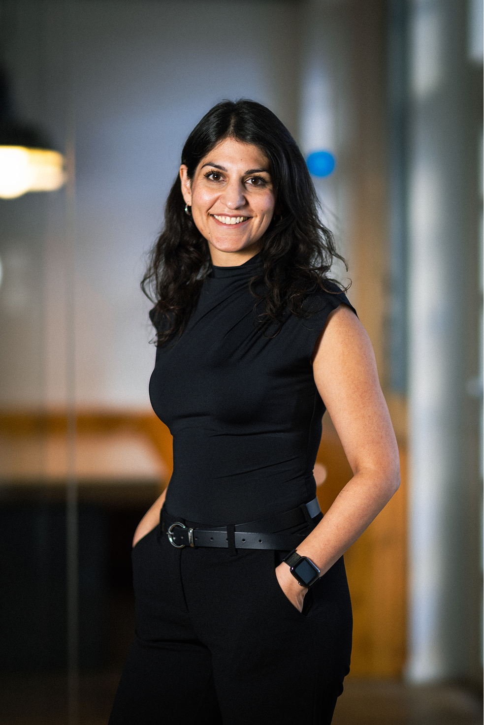 Zamina Ahmad mit langen dunklen Haaren trägt schwarzes ärmelloses Oberteil und schwarze Hose, lächelnd mit Händen in den Taschen.