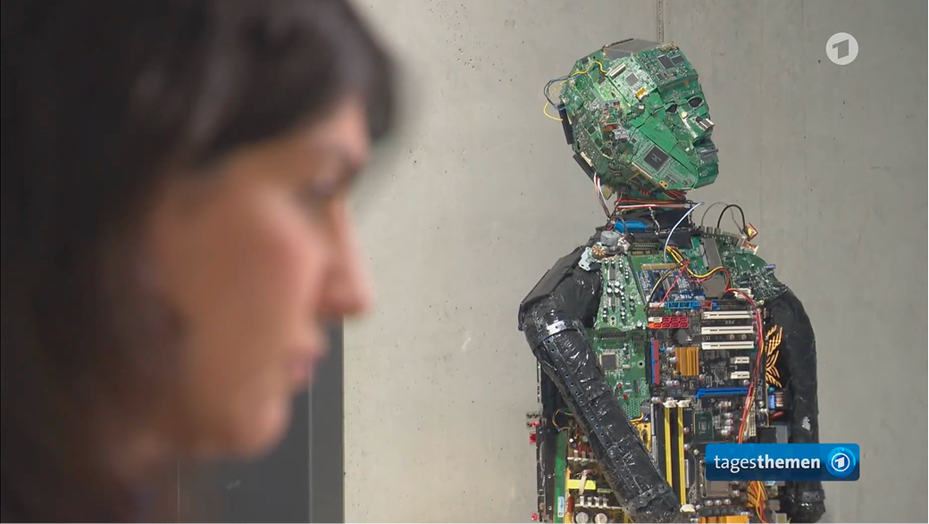 Zamina Ahmad im Vordergrund blickt auf eine Skulptur aus Computerplatinen und elektronischen Bauteilen, die einen menschlichen Torso mit Kopf bildet.