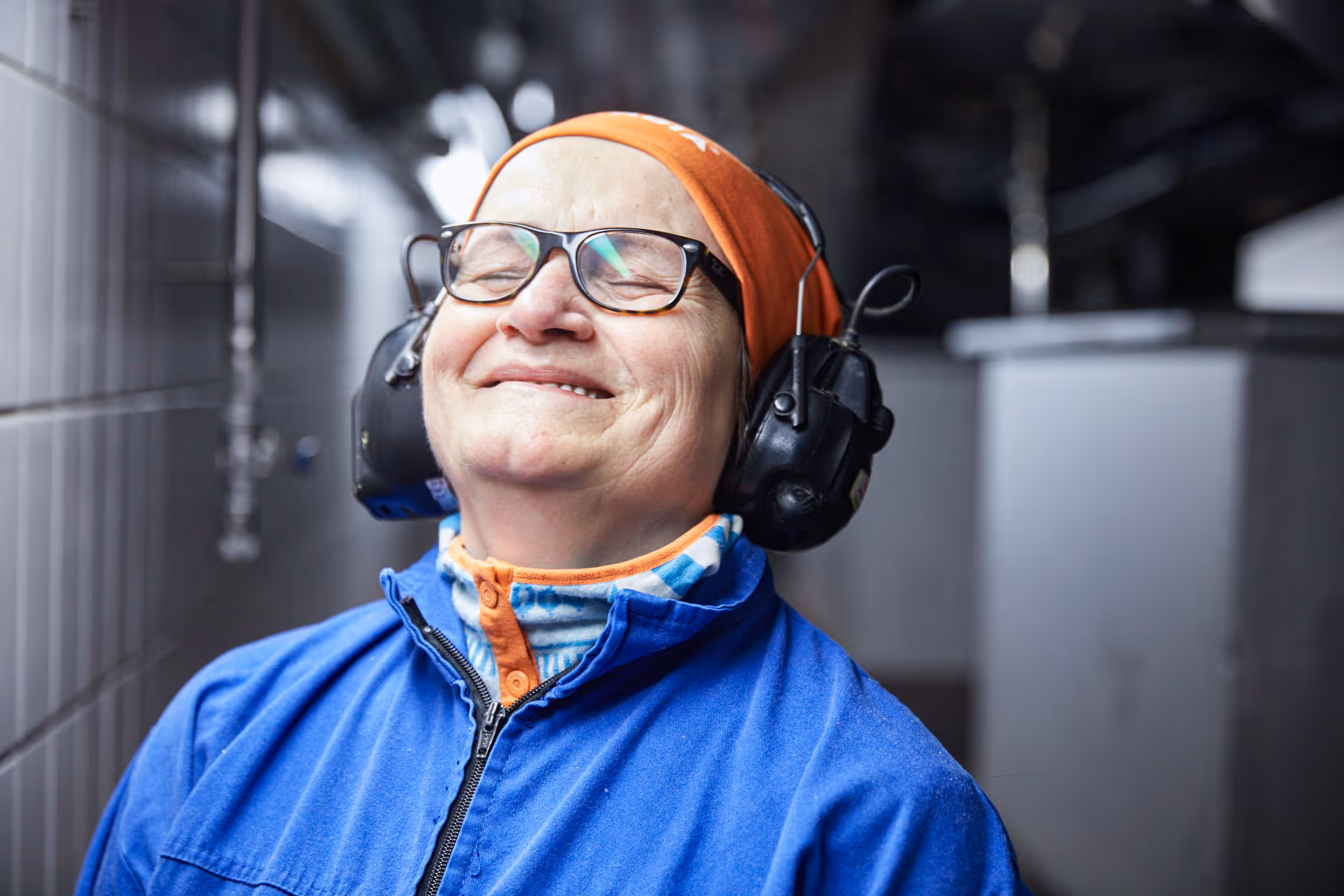 Smiling older woman wearing glasses, blue work jacket, orange headband, and black protective earmuffs in an industrial setting.