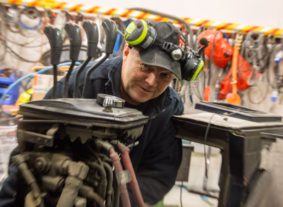 Man wearing a black cap with green headphones working on machinery with control levers in an industrial workshop.