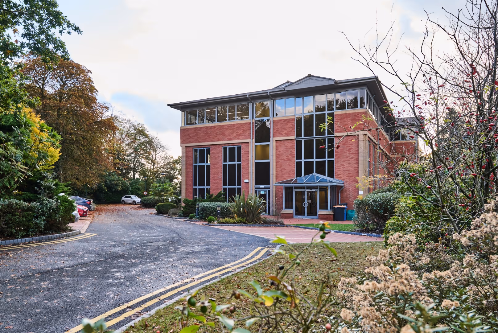 Three-story red brick office building with large windows, surrounded by trees and shrubs, with a driveway and parked cars.
