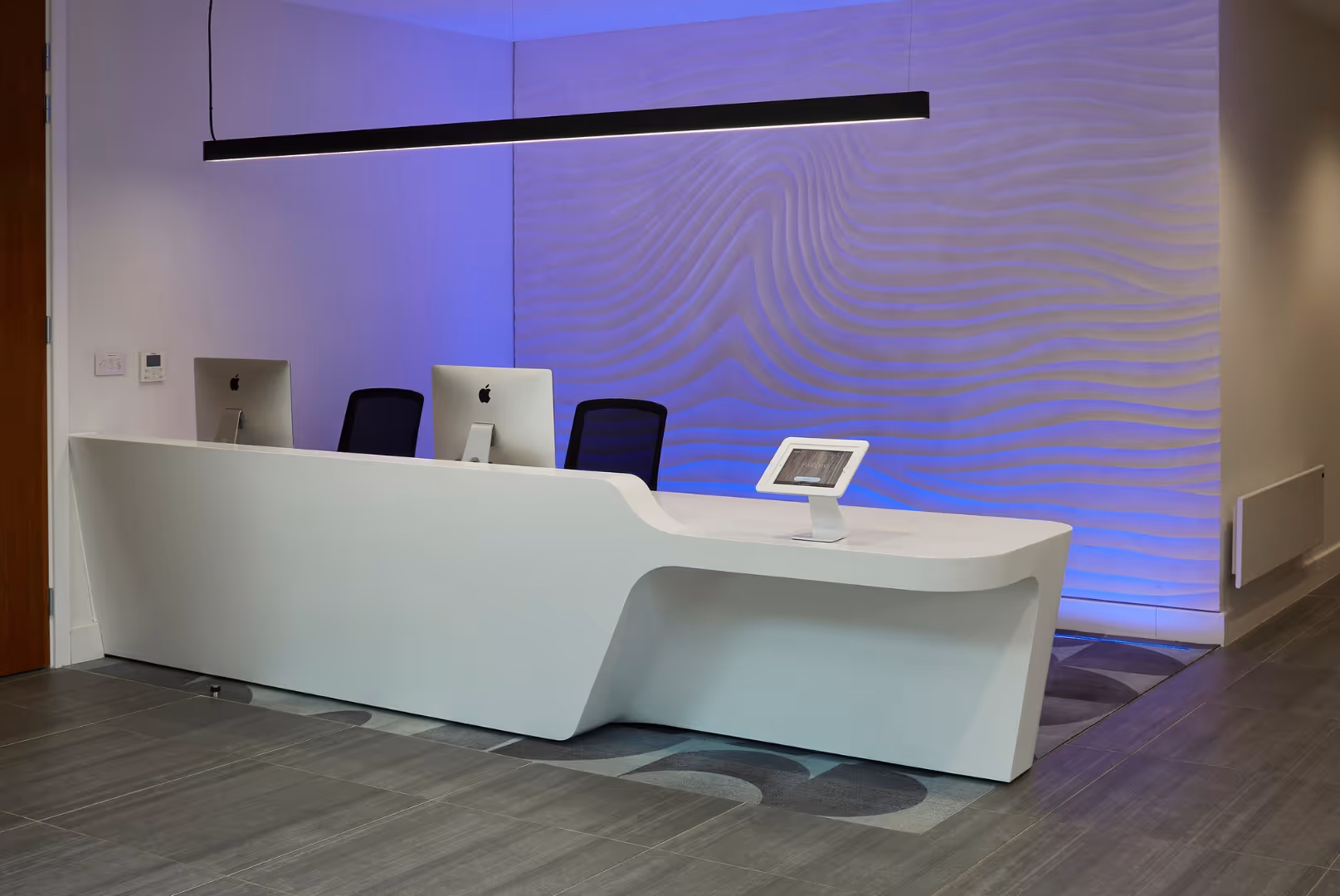 Modern white reception desk with two iMac computers, two black chairs, a tablet, and textured wall illuminated with blue lighting.