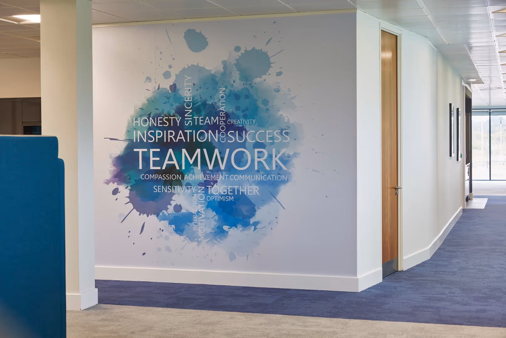 Office hallway wall mural with blue paint splashes and words related to teamwork like 'TEAMWORK', 'INSPIRATION', 'SUCCESS', and 'HONESTY'.