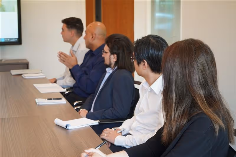 Five professionals attentively watching a presentation in a conference room with notebooks and papers on the table.