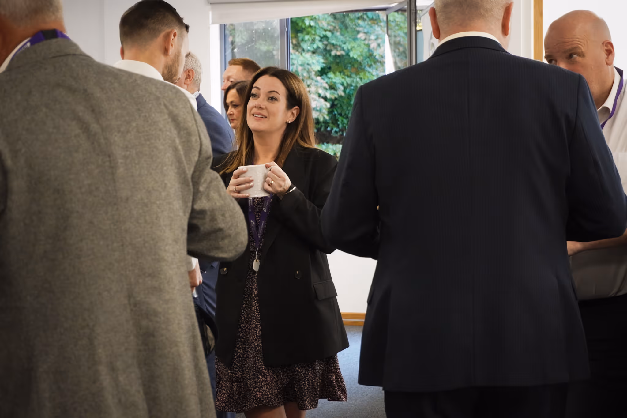 Group of professionally dressed people socializing indoors, one woman holding a cup and engaging in conversation.