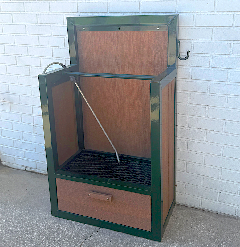 Green and brown outdoor metal and plastic wood show spike cleaner with a metal grate and a metal rod inside, placed on concrete against a white brick wall.