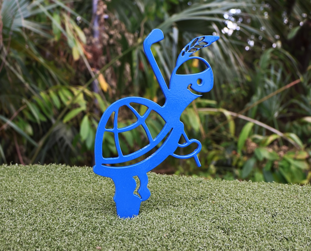 Blue metal tee marker shaped like a smiling turtle holding a golf club and a feather, standing on artificial grass with a blurred green foliage background.