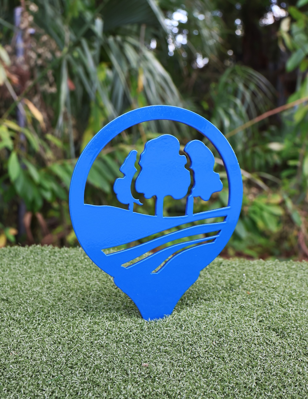 Blue metal tee marker with cutouts of three trees and rolling hills on artificial grass.