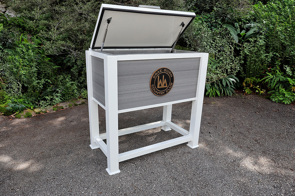 White and grey outdoor cooler with an open lid featuring the Brae Burn Country Club emblem, placed on a paved area surrounded by lush greenery.