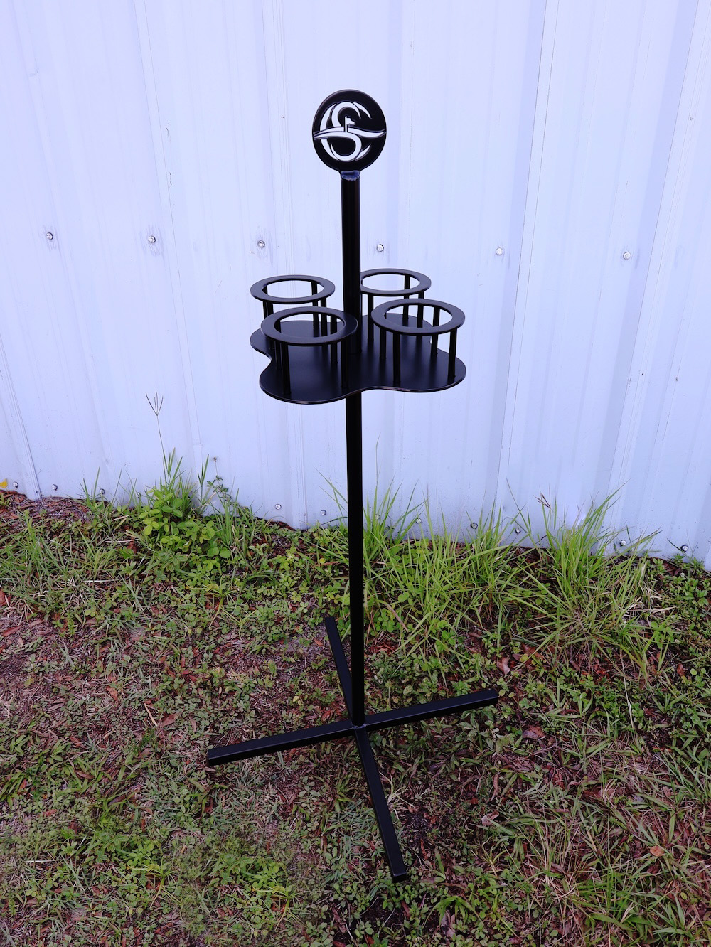 Black metal drink stand with four circular holders and a logo on top, placed outdoors on grass against a white corrugated metal wall.