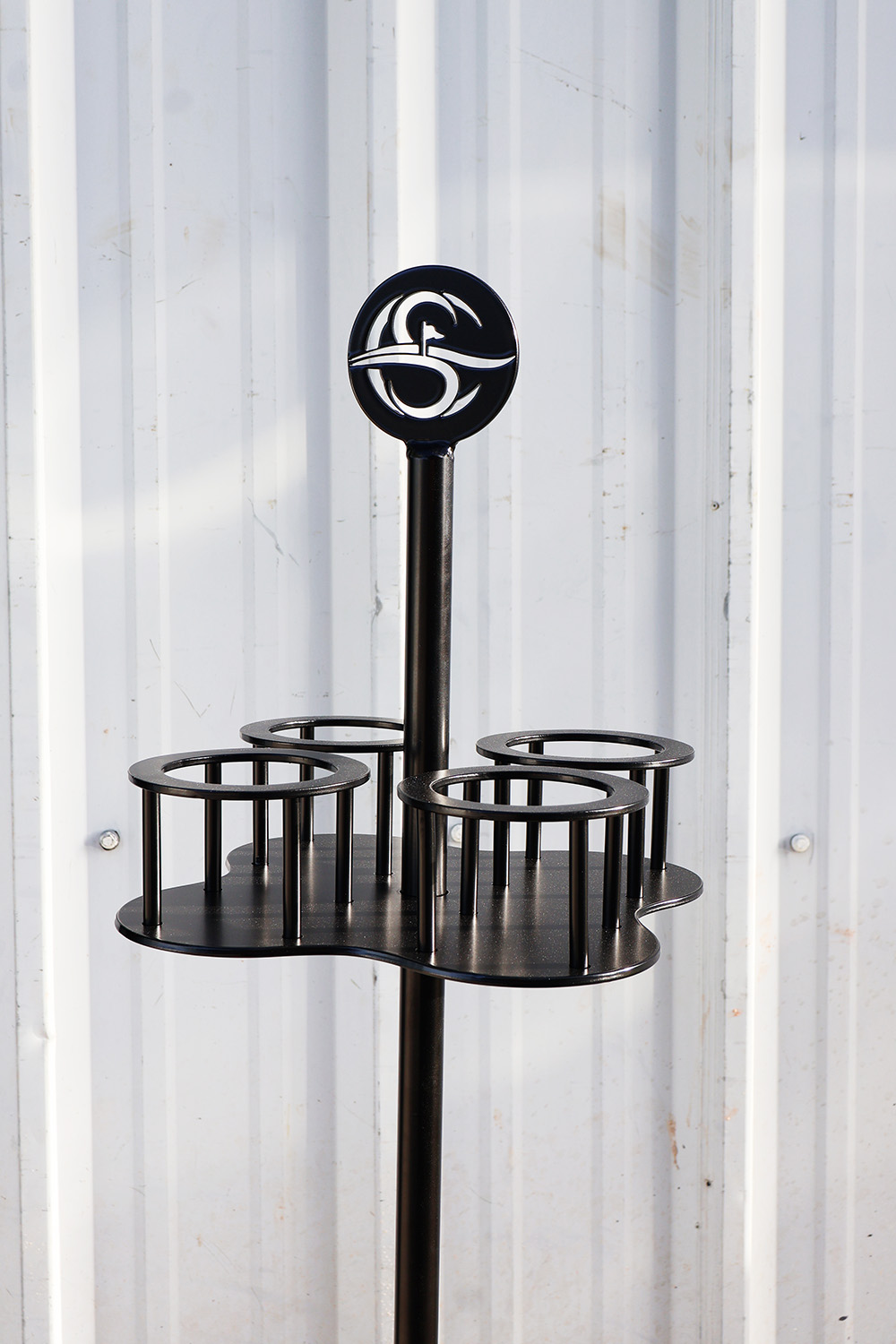 Black metal cup holder with four circular compartments and custom logo mounted on a vertical pole against a white corrugated metal background.