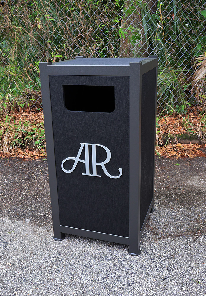 Black outdoor trash can with aluminum frame and AR logo on the front, placed on pavement near a chain-link fence with greenery.