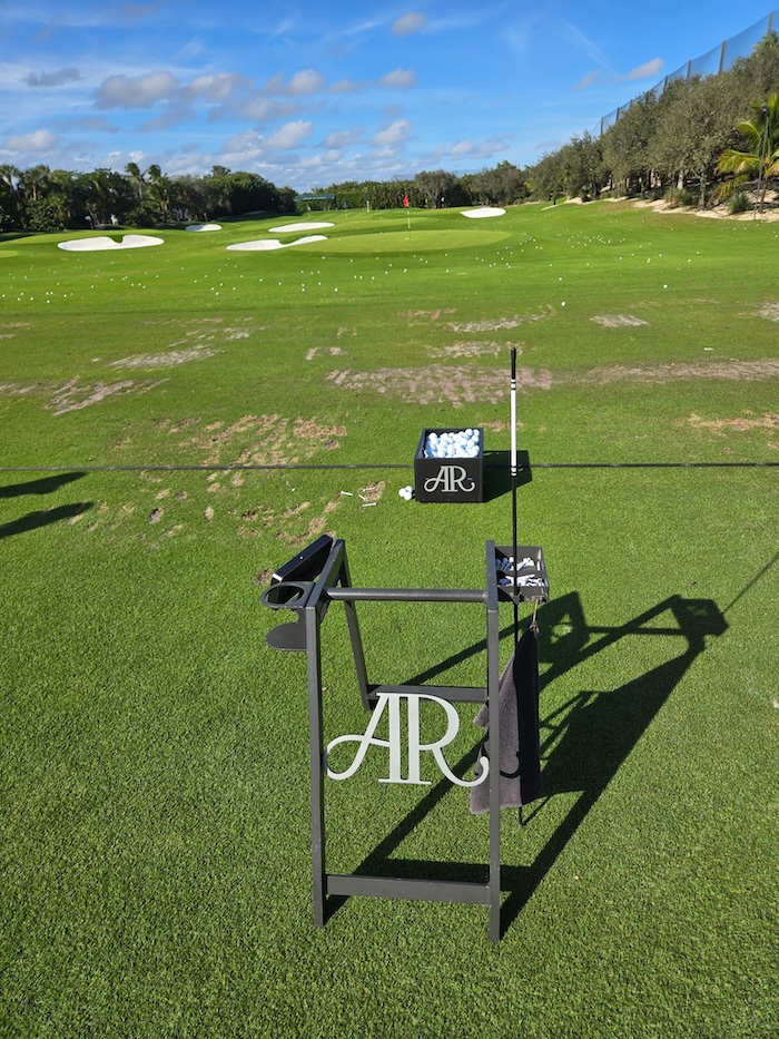 Golf practice range with a ball crate holding golf balls and a bag stand, green grass, sand bunkers, and a clear blue sky.