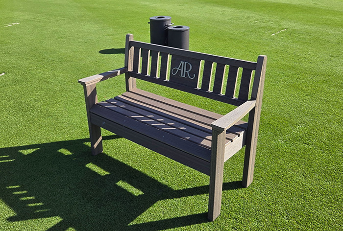 Wooden bench with 'AR' initials on the backrest placed on a green grassy surface with two black club cleaners behind it.