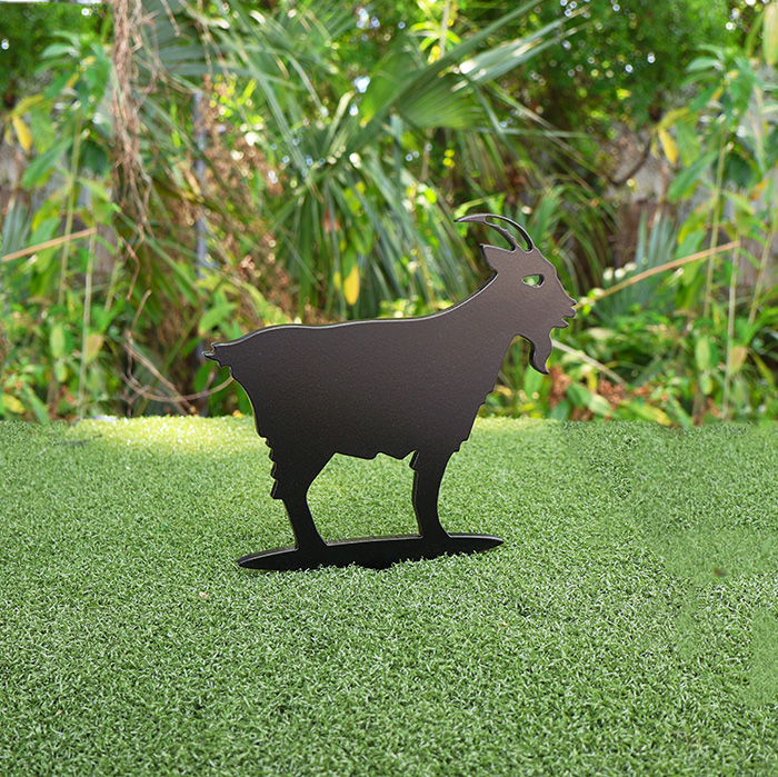 Black metal silhouette of a goat standing on artificial grass with tropical plants in the background.