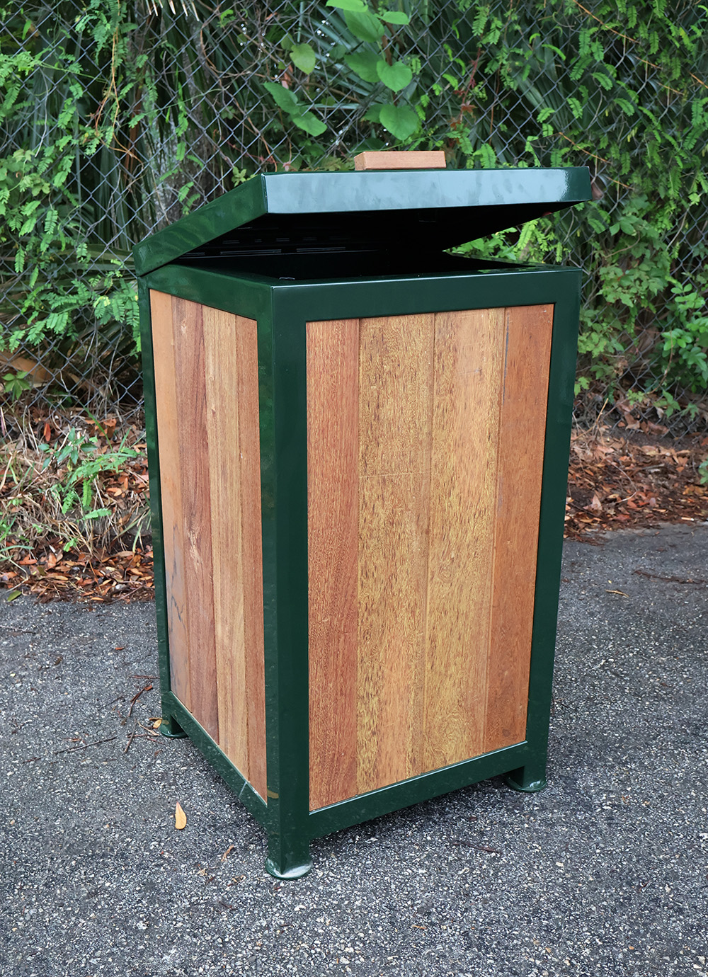 Outdoor trash bin with wood panels and dark green metal frame, lid partially open by chain link fence.