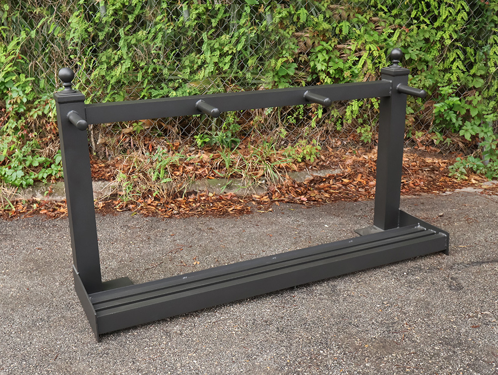 Black metal golf bag stand with three pegs and slotted base on pavement near green foliage