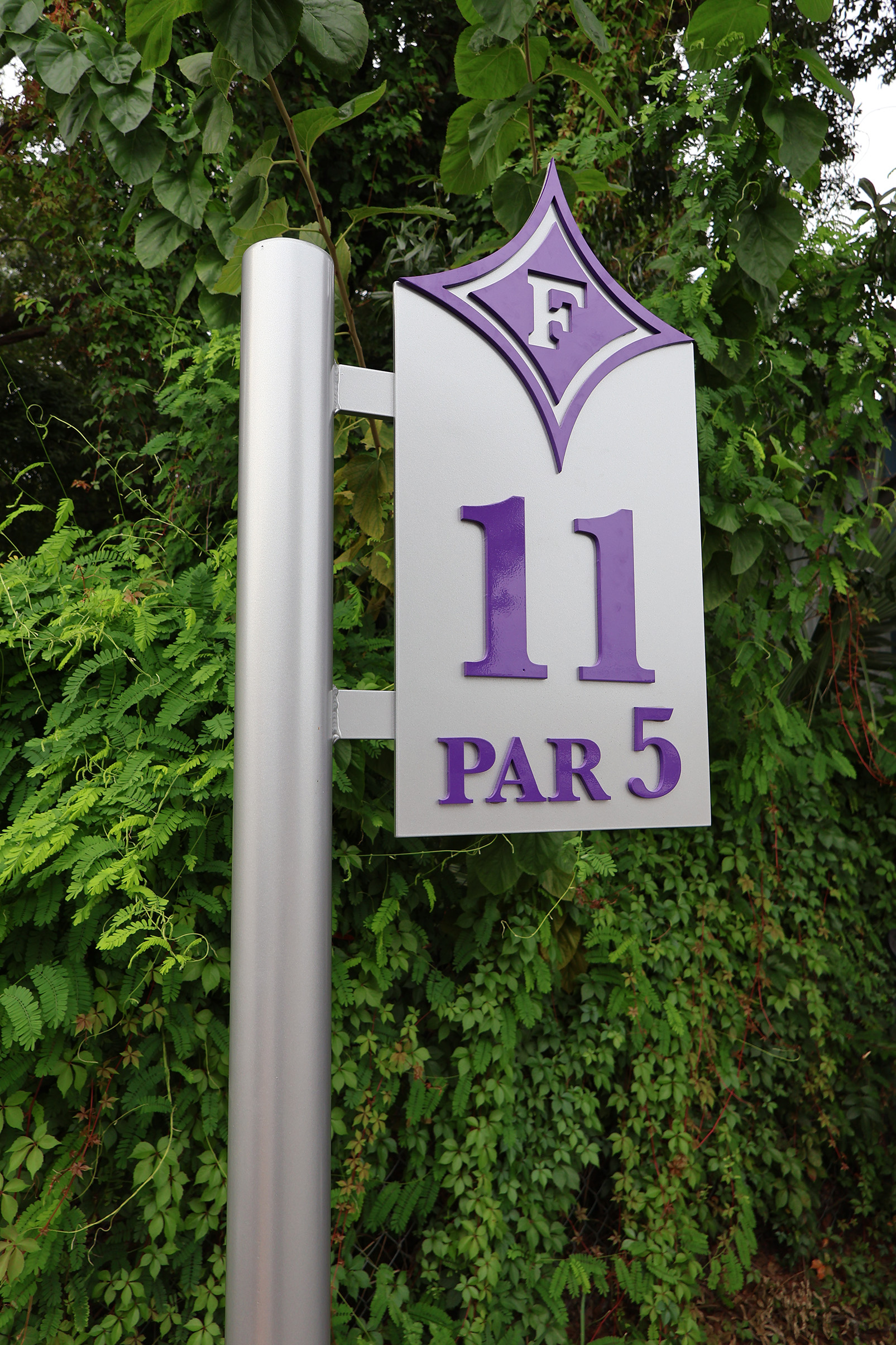Metal sign on silver post with purple F, number 11, and PAR 5 against green foliage.