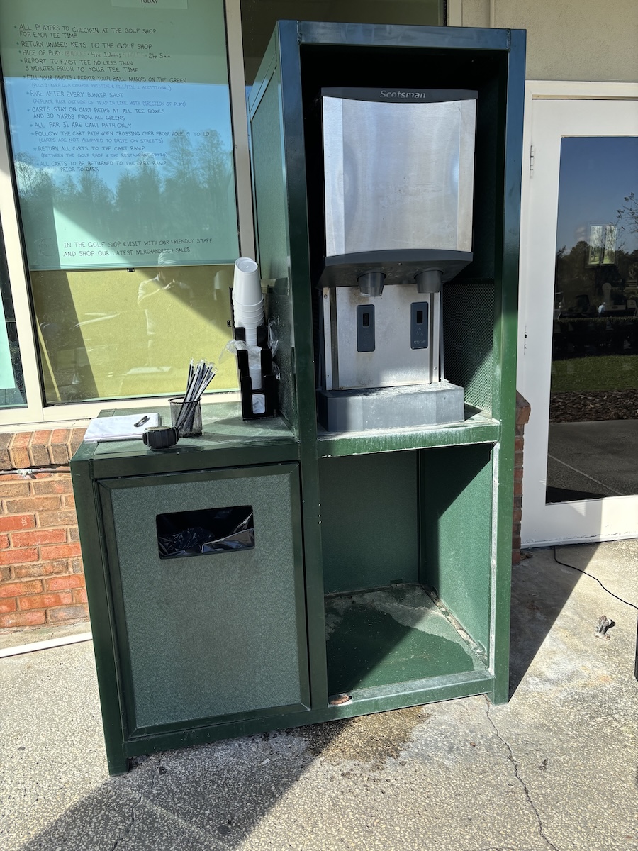 Green outdoor water station with a Scotsman dispenser, paper cups stacked beside it, a small container of straws, and a garbage bin underneath, positioned next to a building with a glass door and window displaying golf shop rules.