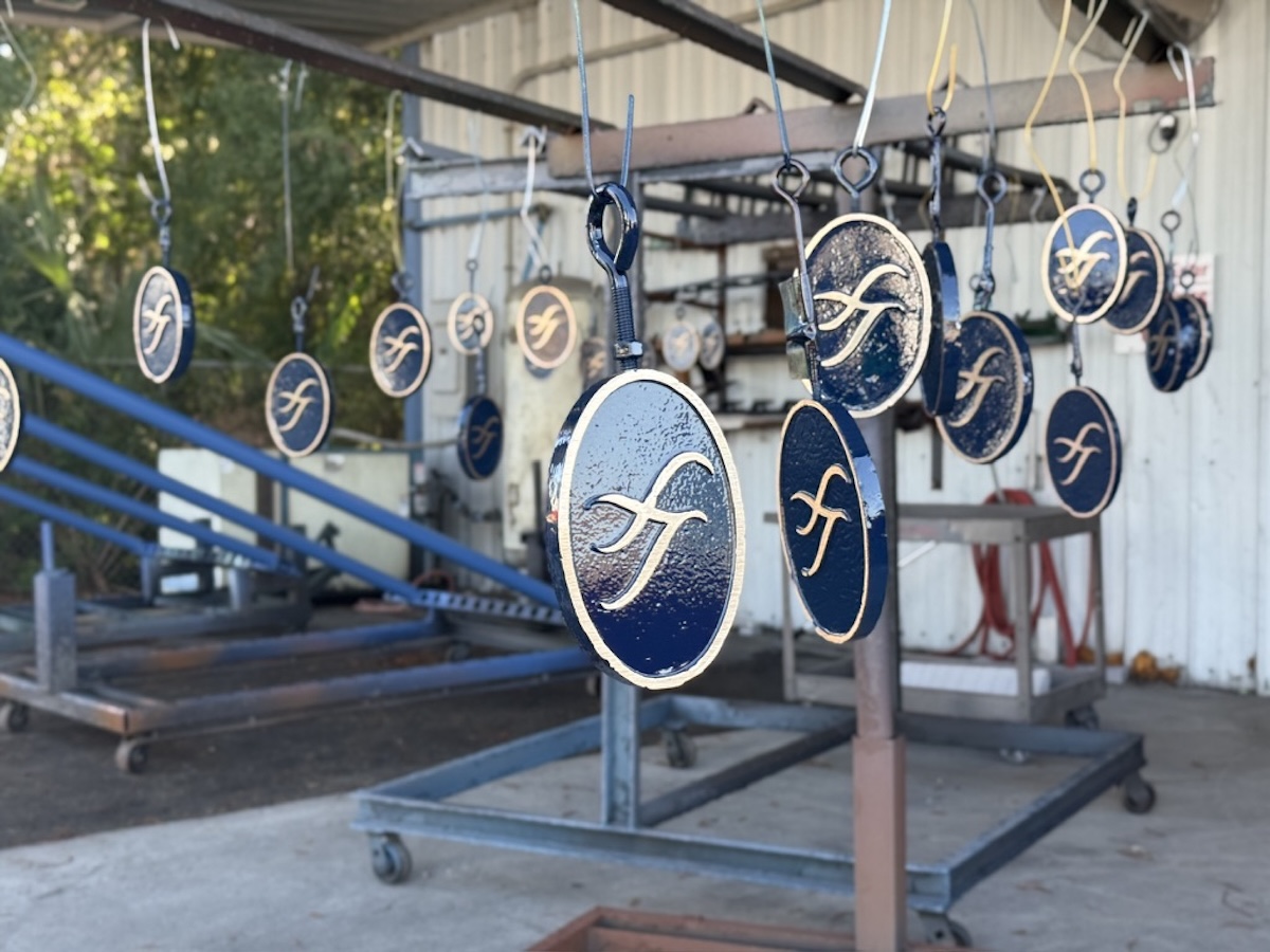 Multiple refurbished cast metal tee markers painted blue with a gold letter 'lj' embossed on them, hanging from wires inside a workshop with metal frames and outdoor natural light.