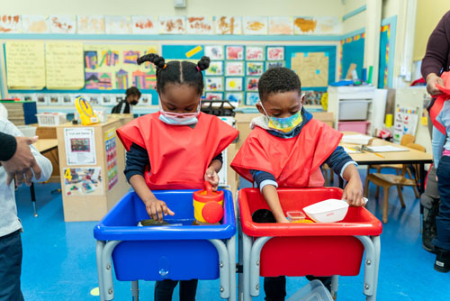 Two masked kindergartners explore sensory bins wearing red smocks in classroom