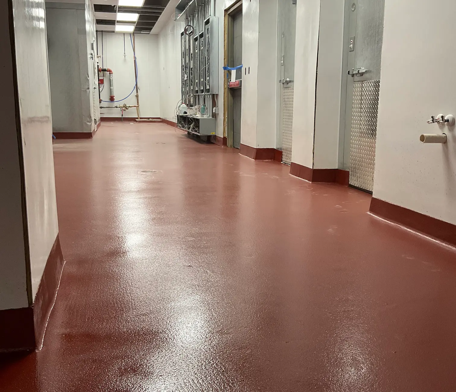 Long industrial hallway with smooth reddish-brown floor and white walls with metal doors and pipes.