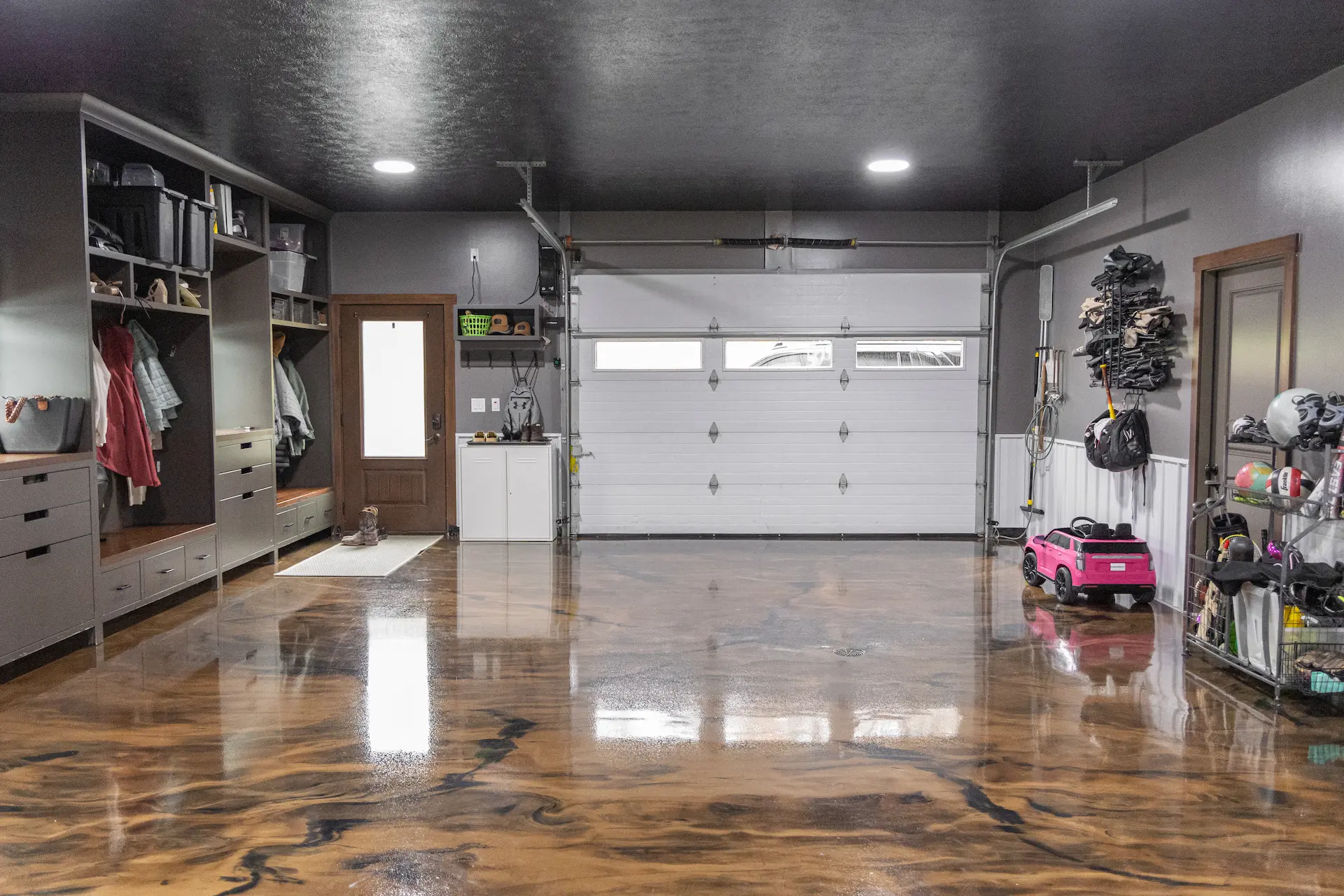 Spacious residential garage with polished epoxy-coated floor featuring a marbled brown and black pattern, white sectional garage door, gray walls, built-in storage cabinets, shelves with shoes and clothes on the left, and sports equipment including a pink toy car and helmets on the right.