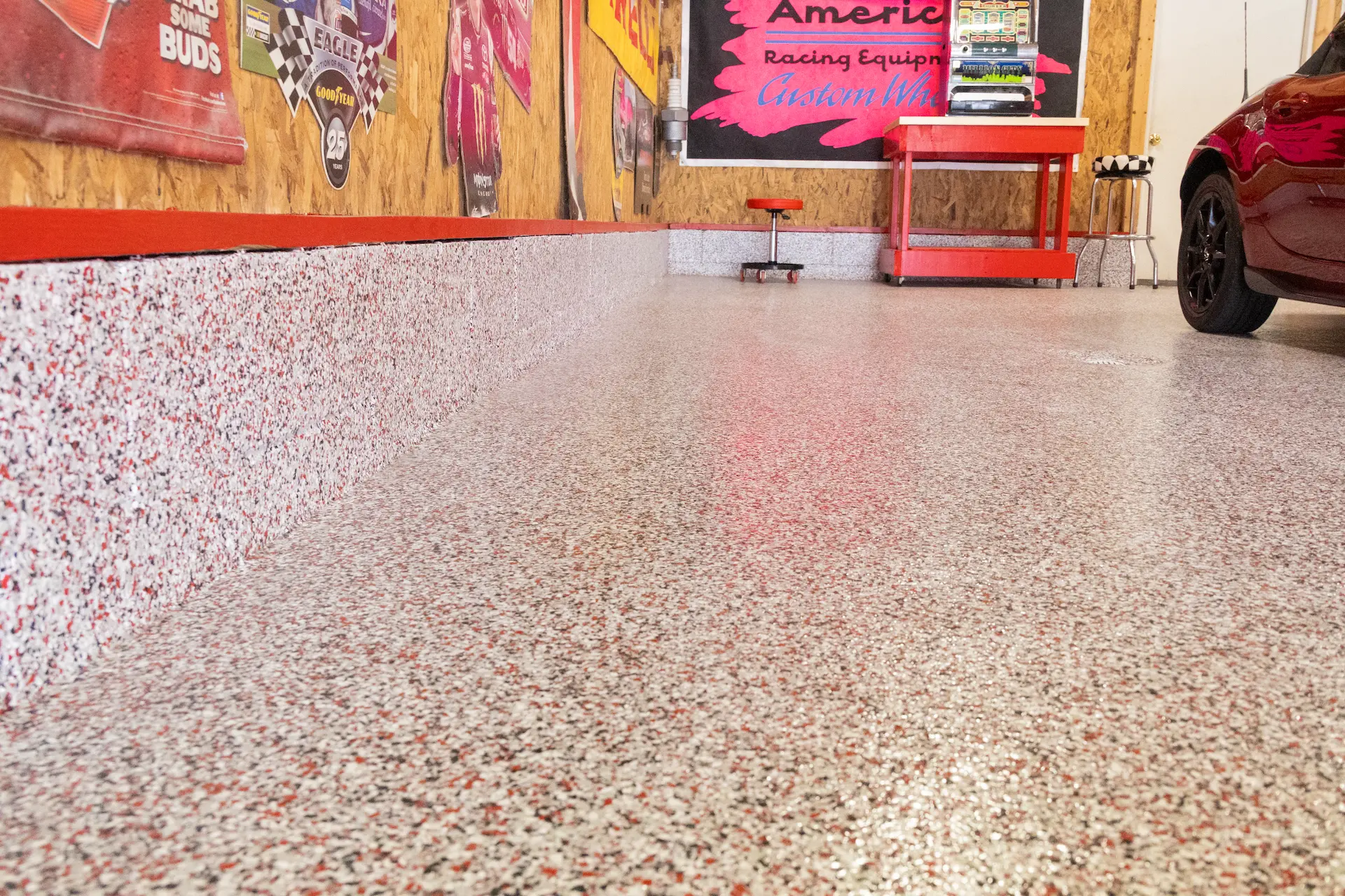 Epoxy floor coating with red, white, and black flakes covering the floor and baseboard of a garage, with a red tool stool, a red workbench, and a partial view of a red car on the right.