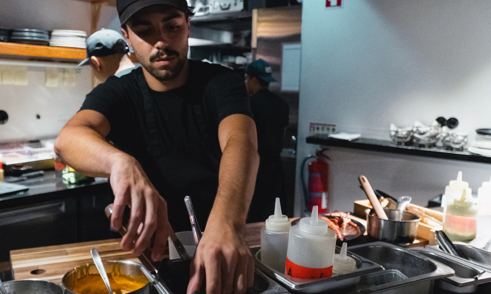 Male chef in a black shirt and cap preparing food in a professional kitchen with various condiments and utensils on the counter.