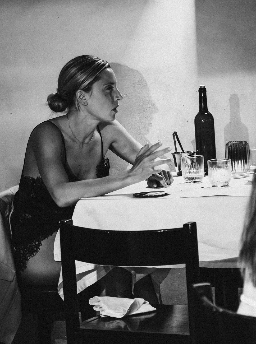 Black and white photo of a woman in a black dress sitting at a table with glasses and a bottle, gesturing with her hand.