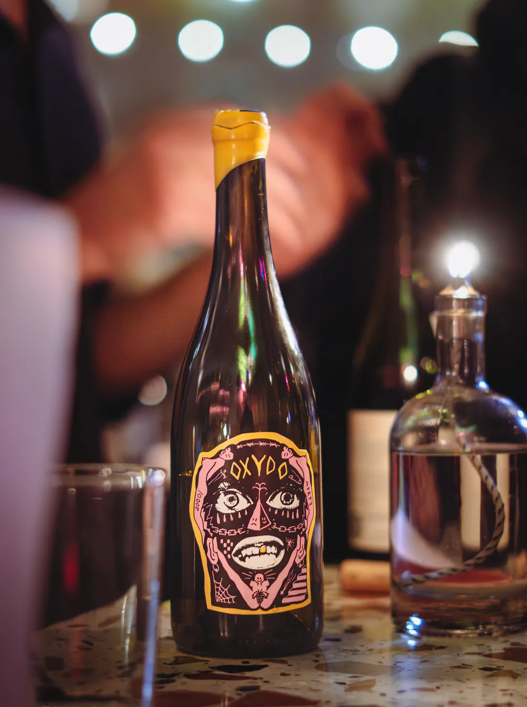 Dark bottle with a yellow cap and a colorful label featuring an abstract face with large eyes and teeth on a bar counter.