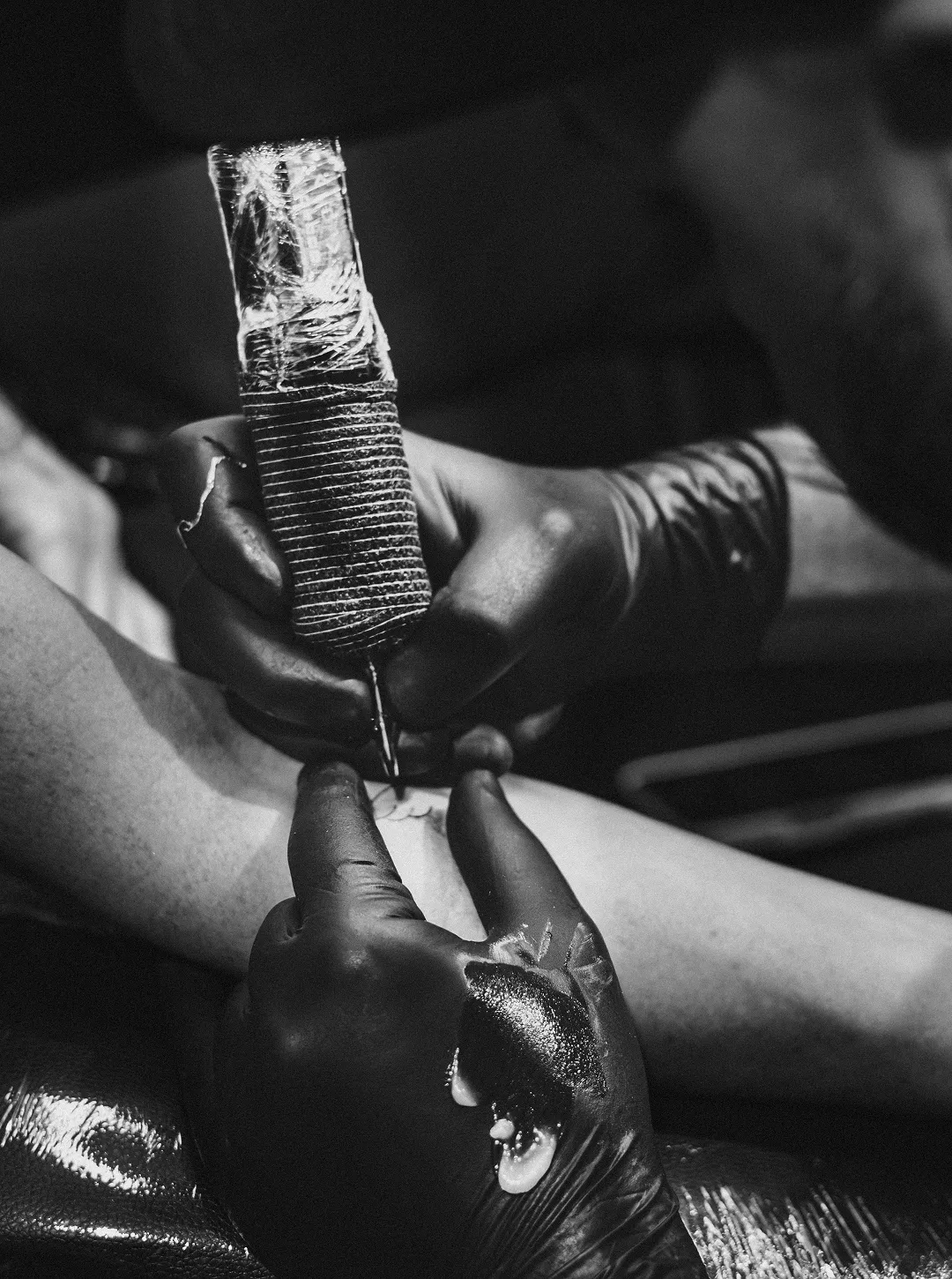 Close-up black and white photo of a tattoo artist's gloved hands working on a tattoo on a person's arm.