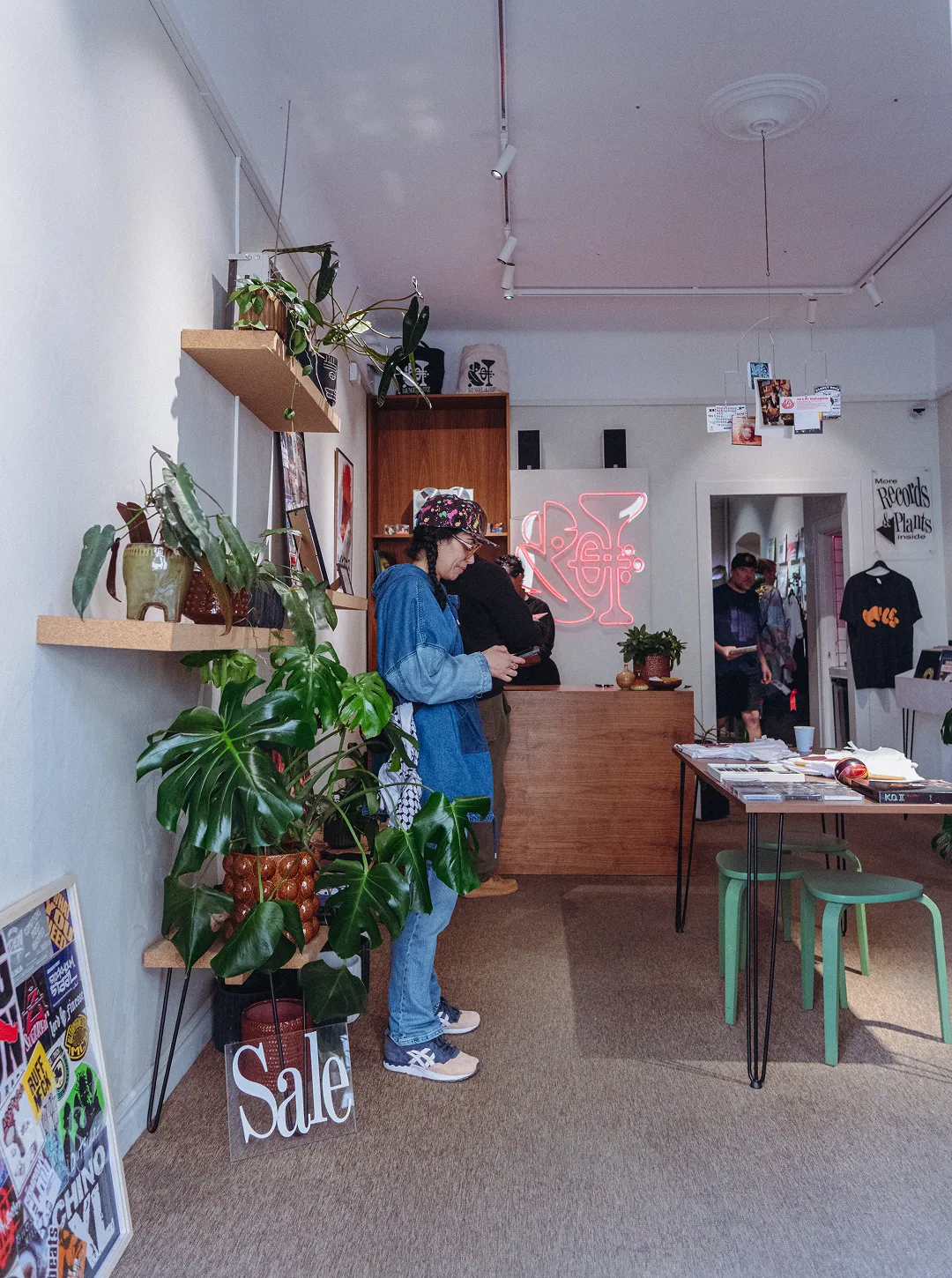Interior of a boutique shop with plants, shelves, a wooden counter, a person in a colorful cap looking at a phone, and a neon sign on the back wall.