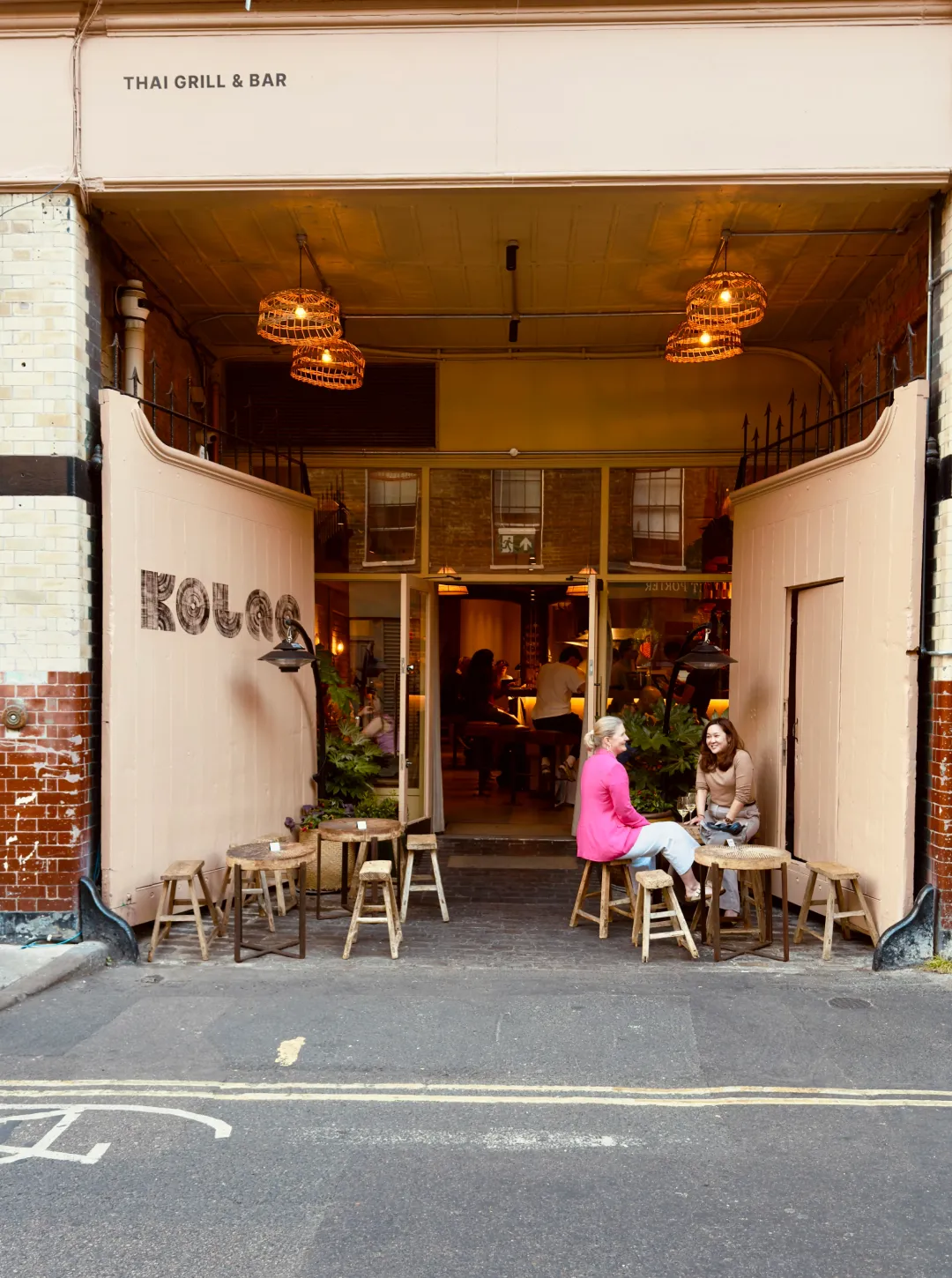 Two women sitting at small wooden tables outside a Thai grill and bar with open doors showing people inside the warmly lit restaurant.