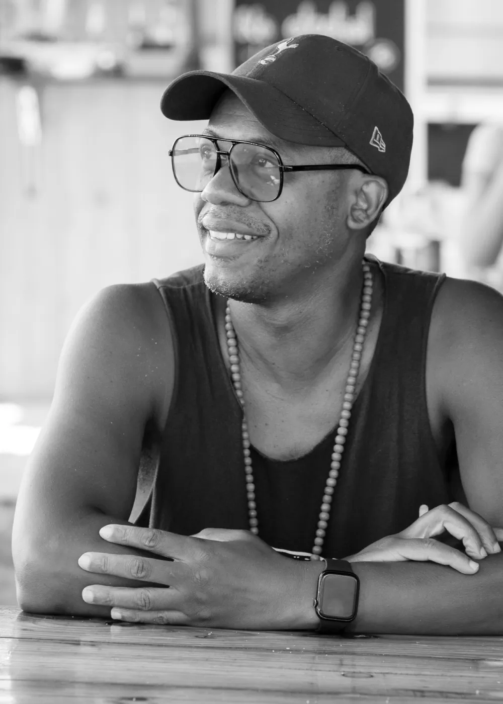 Rod Lewis smiling and  wearing a cap, large glasses, a beaded necklace, and a smartwatch, sitting at a wooden table.