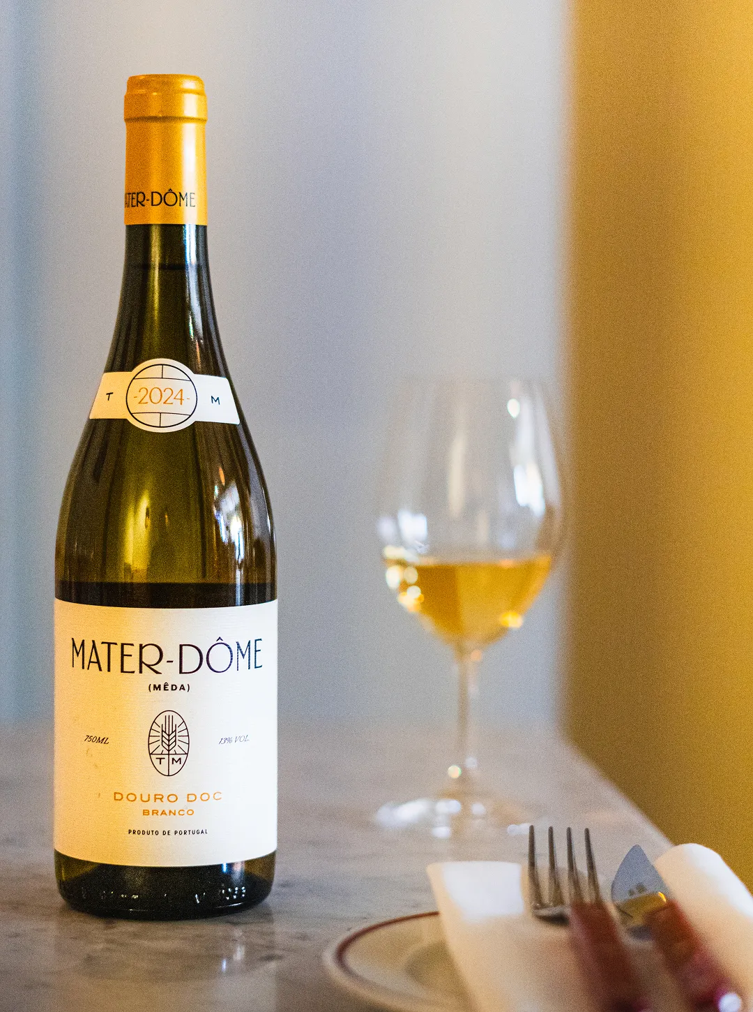 Black and white photo of a woman with hair in a bun sitting at a table with glasses and a bottle, gesturing with her hand.