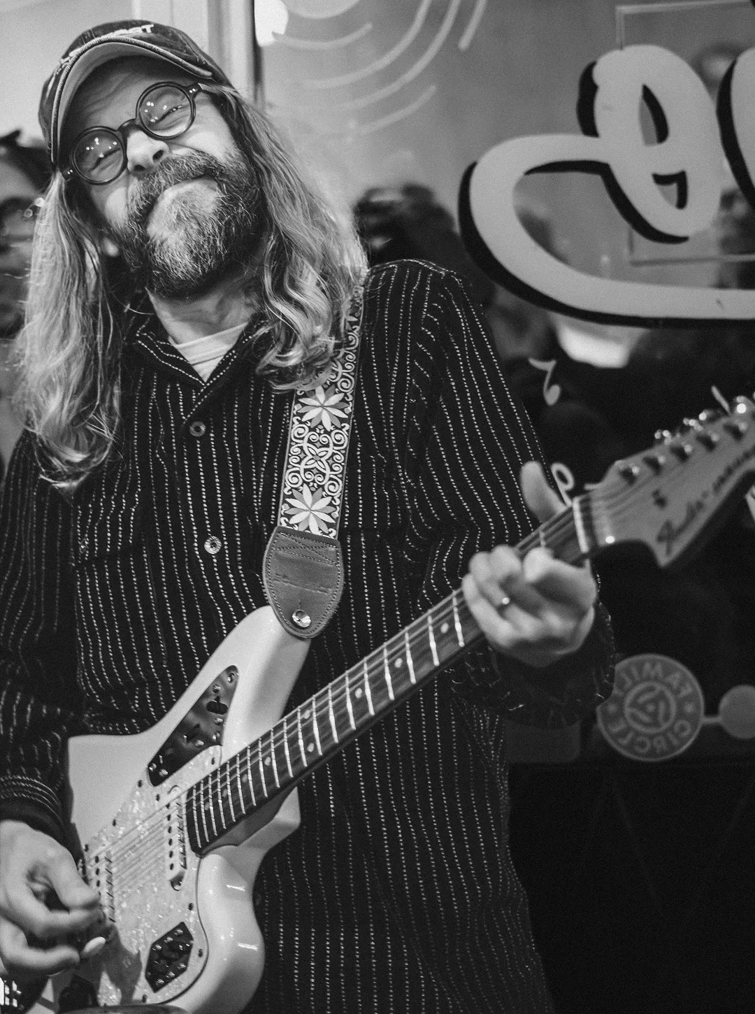 Two people interacting at a wooden counter in a record store, with one person wearing a colorful cap and glasses, and a framed red flower poster on the wall.