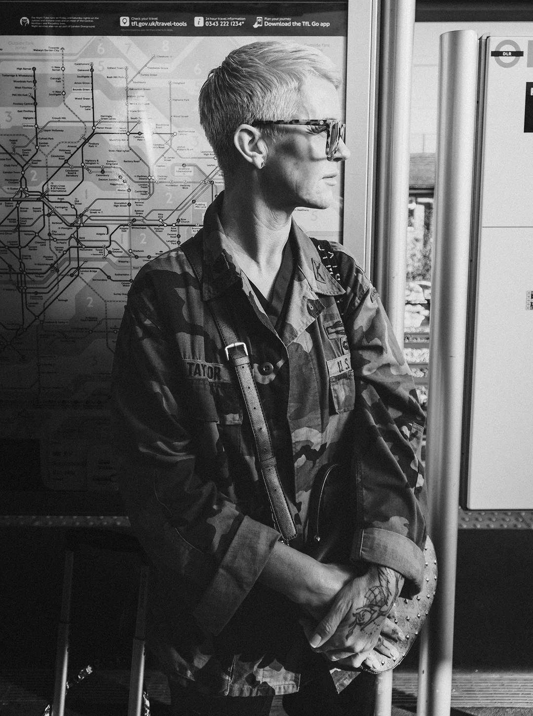 Person with short hair and glasses wearing a camouflage jacket standing near a transport map, holding a studded bag with tattooed hands.