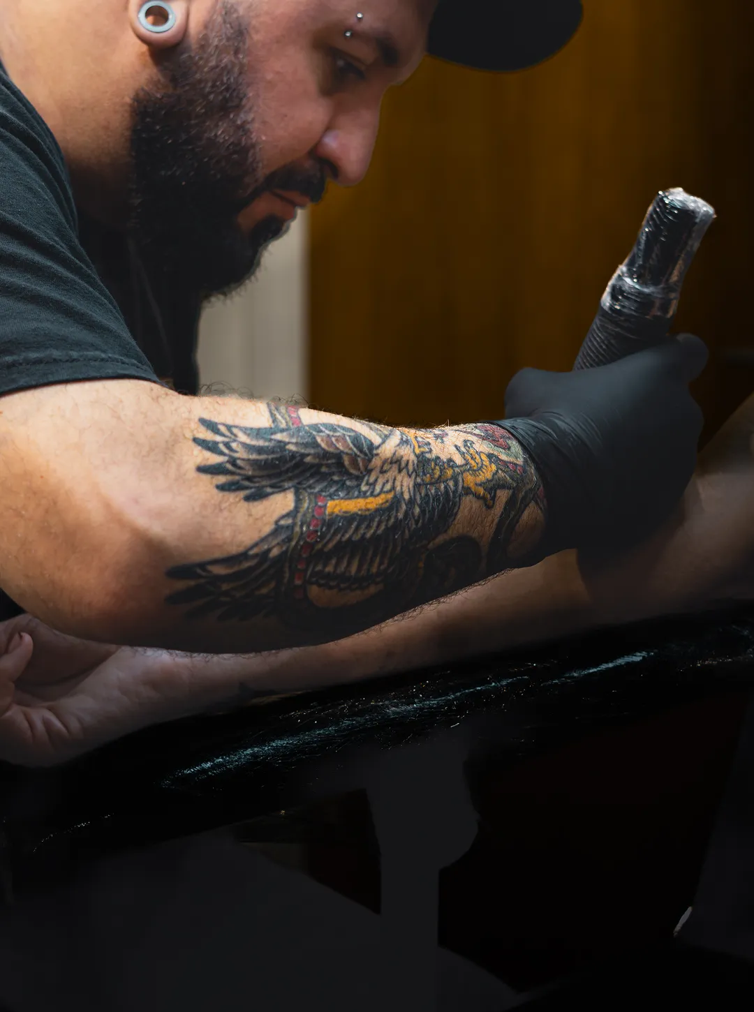 Man with tattoos and a Quadra cap drizzling sauce over a takeout container filled with beans, skewered meat, and pickled vegetables in a kitchen.