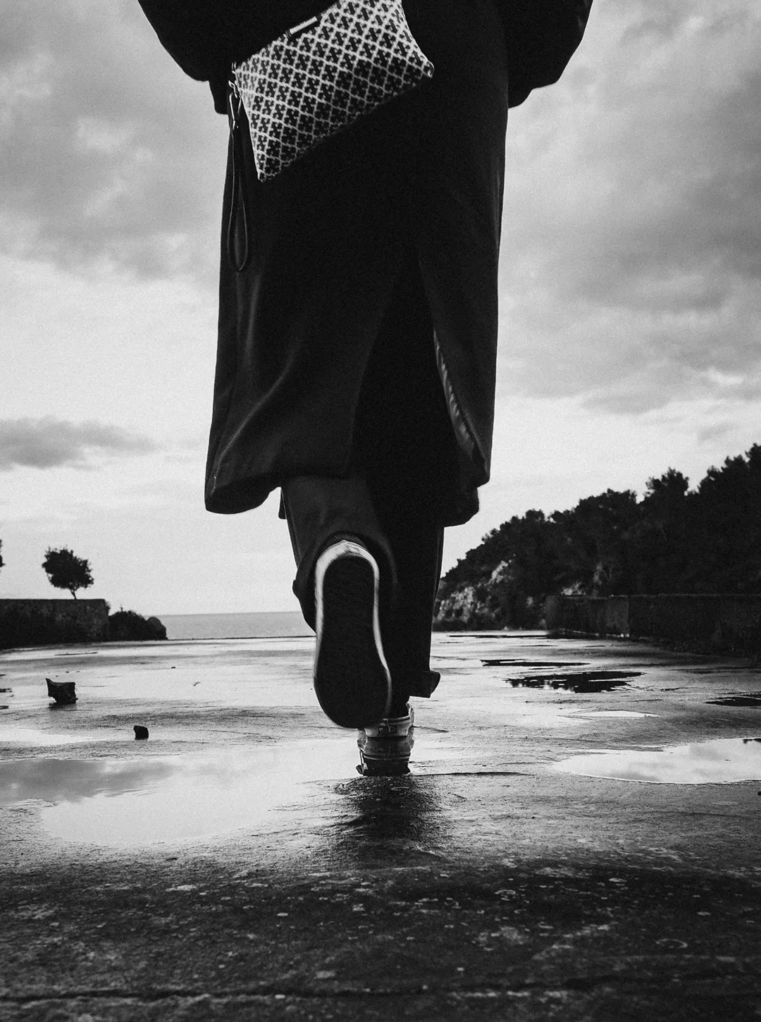 Person walking away on a wet path with puddles, wearing dark clothing and carrying a patterned bag.
