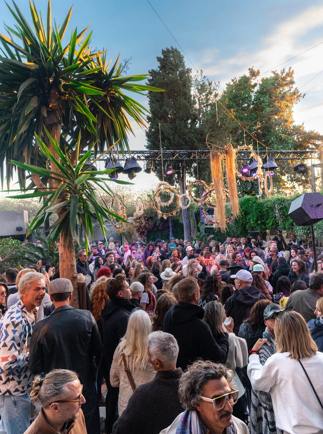 Large outdoor crowd at a lively event under hanging decorations with trees and clear sky in the background.