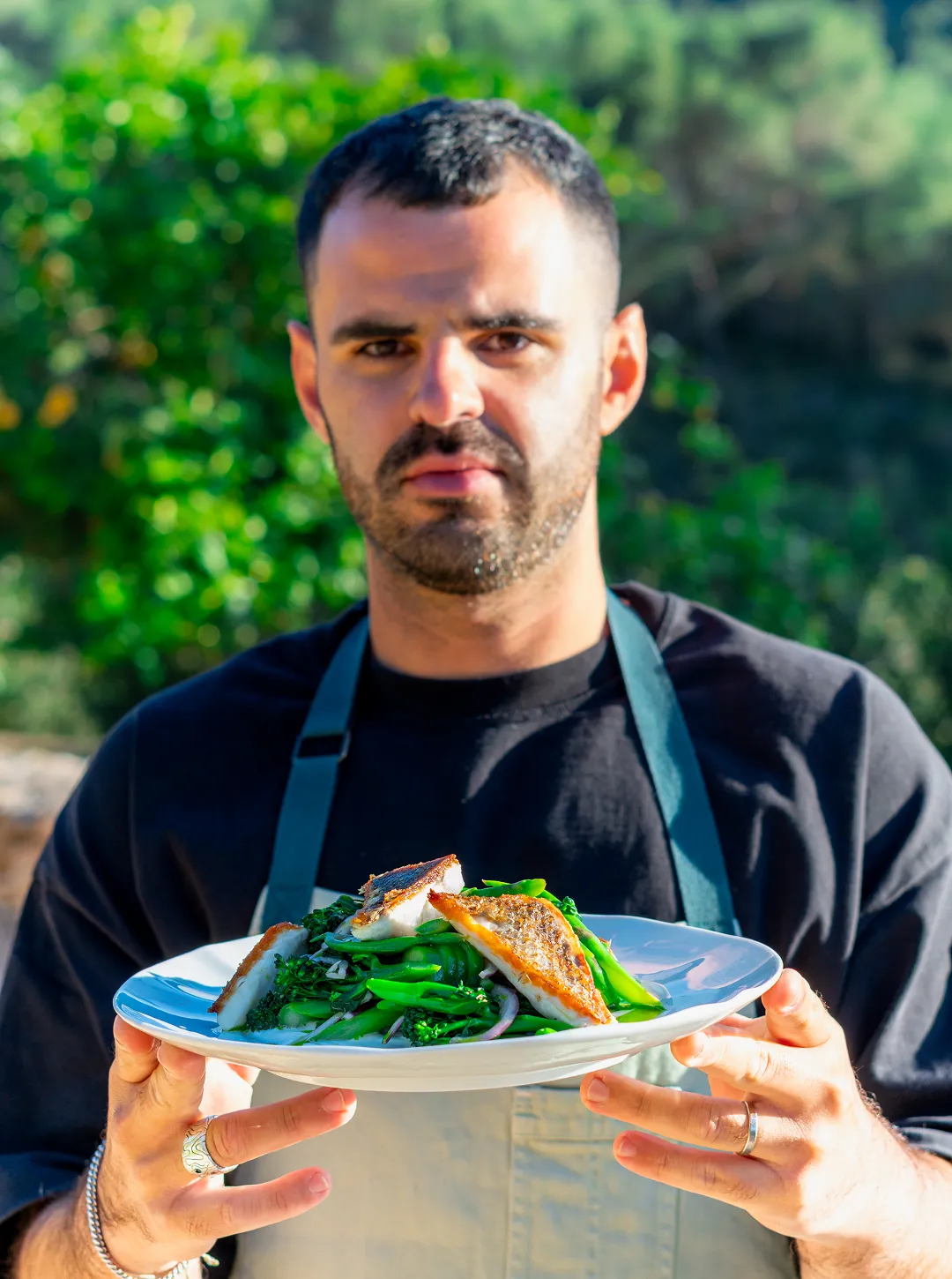 Amit Alif wearing apron holding a plate with cooked fish fillets on a bed of green vegetables outdoors.